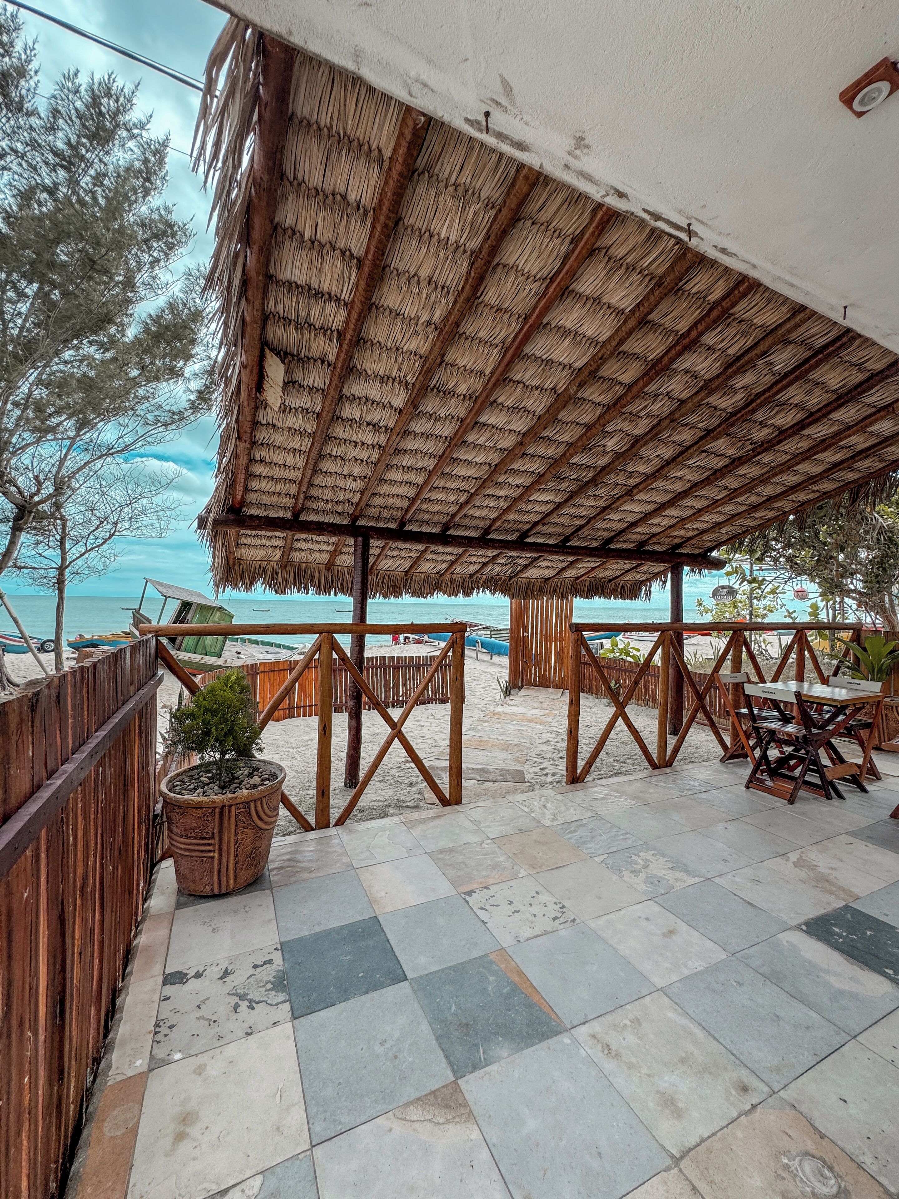 A beachside patio with a thatched roof, wooden furniture, and a view of the sea.