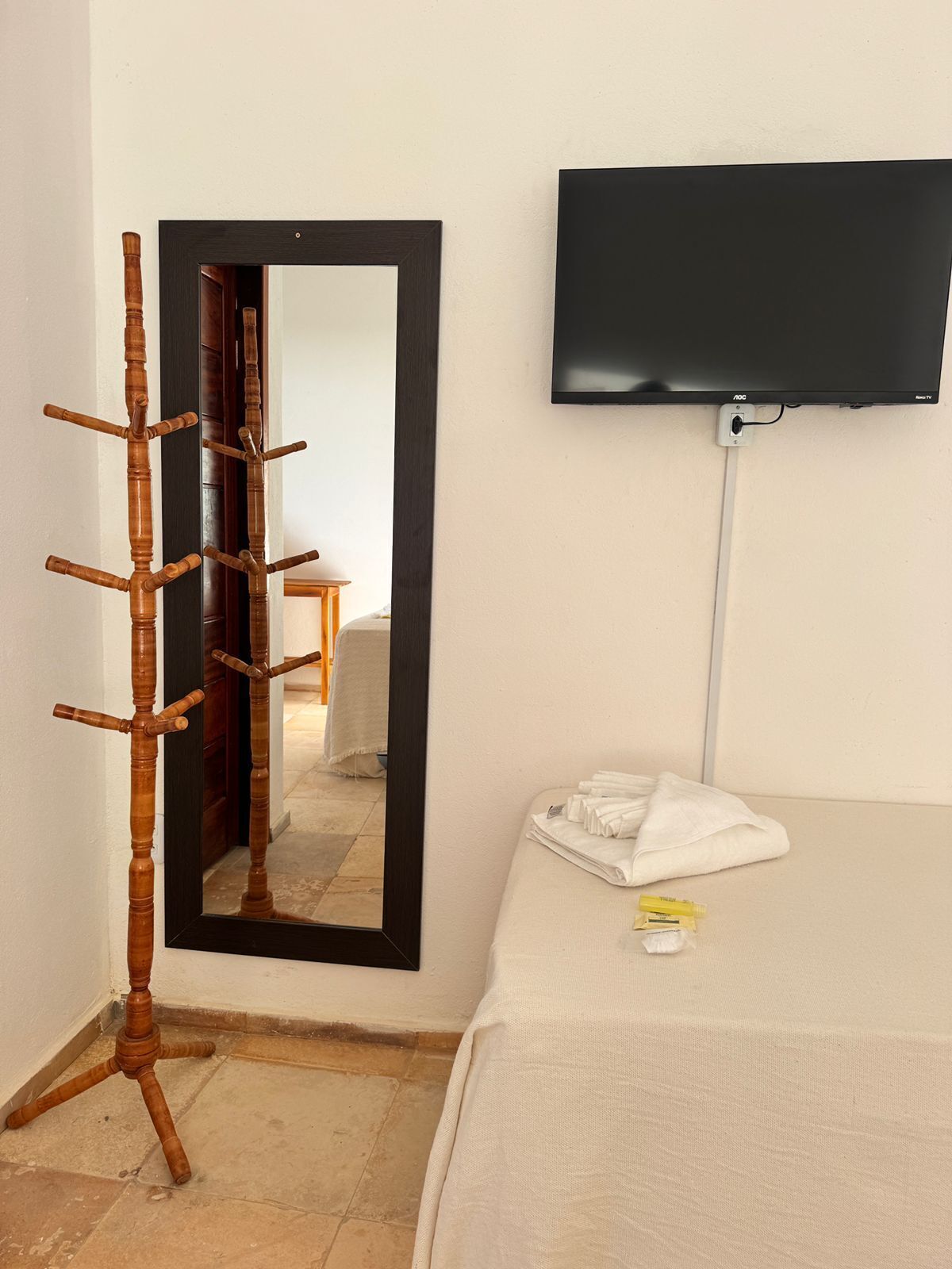 A hotel room with a wooden coat rack, a large mirror, a flat-screen TV mounted on the wall, and a neatly made bed with a white bedsheet.