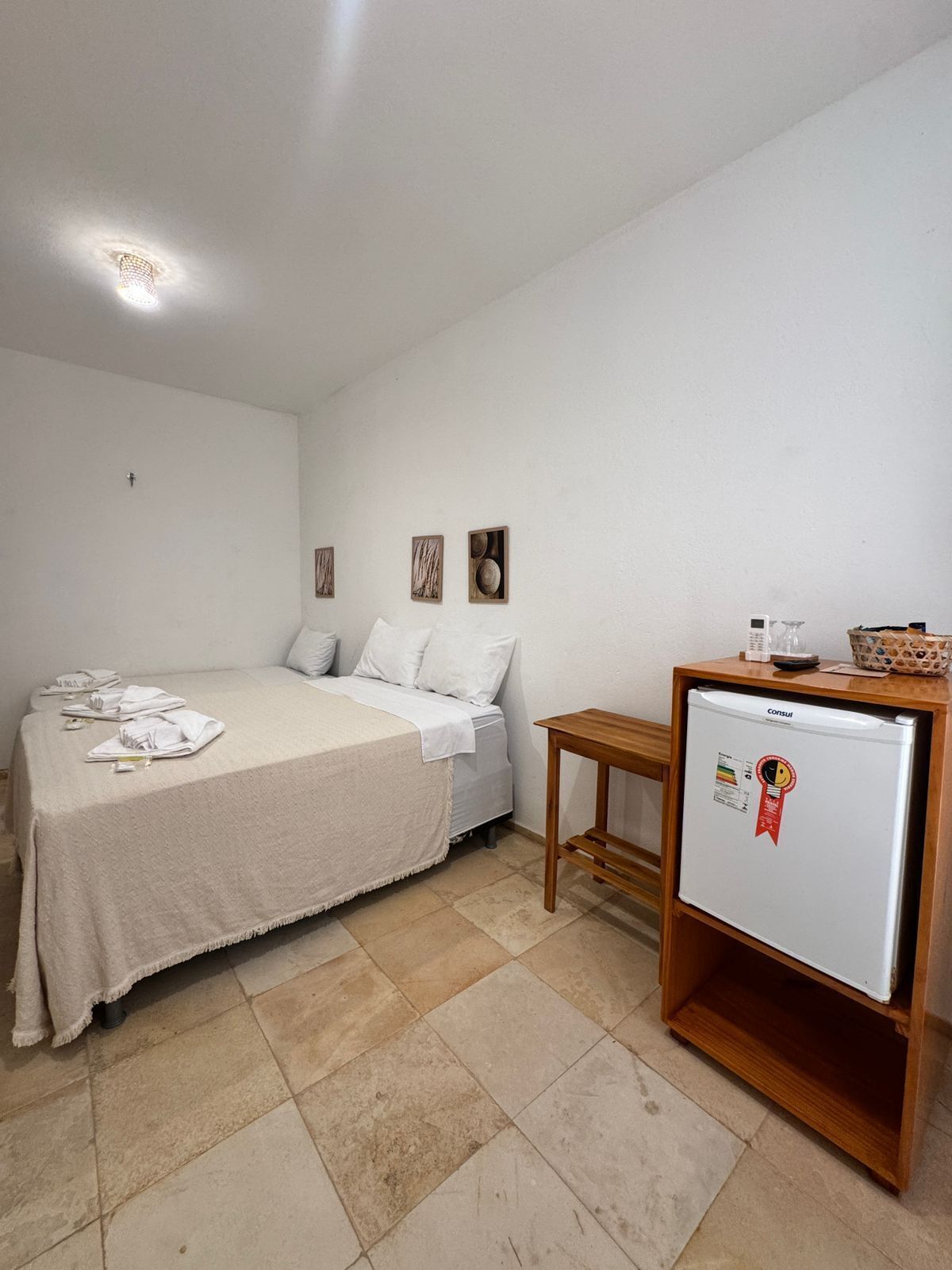 A small, clean room with a bed, a bedside table, a refrigerator, and a wall with three framed pictures.