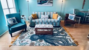 Una sala de estar con un sofá azul, un sillón a juego, un tapete estampado y un cuadro de un barco en la pared.
