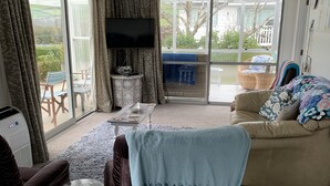 A living room with a sofa, armchair, and a television. There is a sliding glass door leading to an outdoor area with a table and chairs. - A Cosy Beach Cottage Charmer (Riverton)