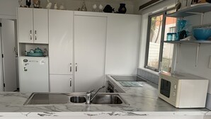A modern kitchen with white cabinets, a double sink, a microwave, and decorative items on shelves. - A Cosy Beach Cottage Charmer (Riverton)