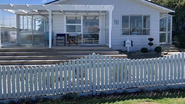 A white cottage with a covered porch, numbered 16, and a white picket fence in front. - A Cosy Beach Cottage Charmer (Riverton)