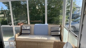 A wicker sofa with cushions and a throw blanket, positioned in a sunlit room with large windows. - A Cosy Beach Cottage Charmer (Riverton)