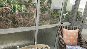 A cozy seating area with woven chairs, a round table with seashells, and a view of a garden through large windows. - A Cosy Beach Cottage Charmer (Riverton)