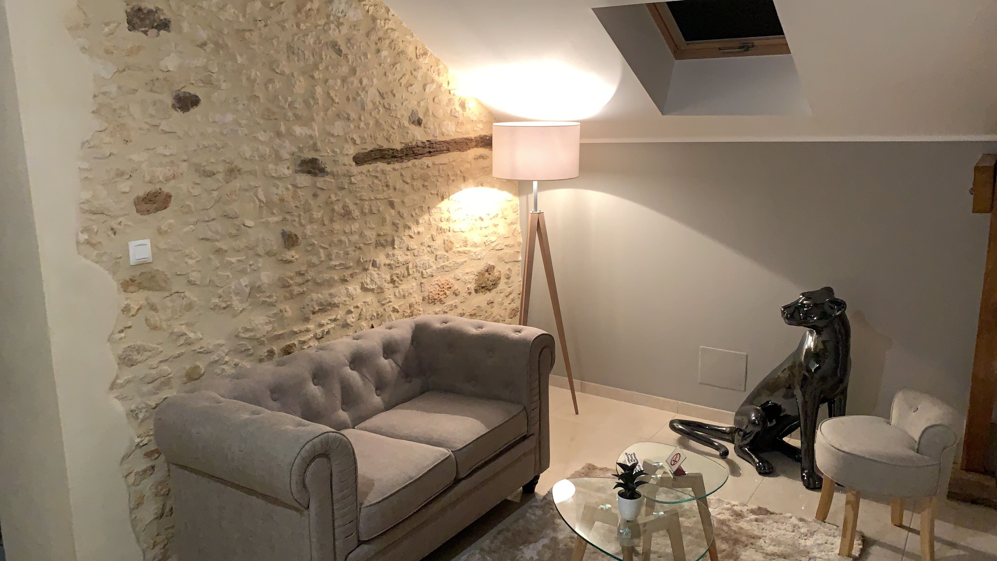 Una sala de estar con pared de piedra, un sofá gris, una lámpara de pie, una mesa de vidrio con una planta y una escultura.