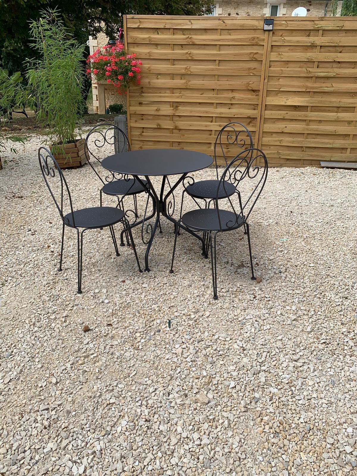 A round metal table with four chairs in a gravel area, surrounded by a wooden fence and greenery.