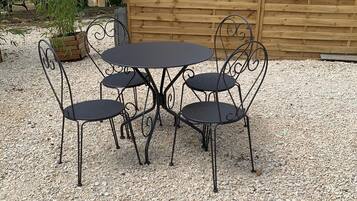 A round metal table with four chairs in a gravel area, surrounded by a wooden fence and greenery.