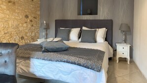 A bedroom with a stone wall, a bed with grey bedding, a nightstand with a lamp, and a small table with a lamp. - La biche au bois  (Montclera)