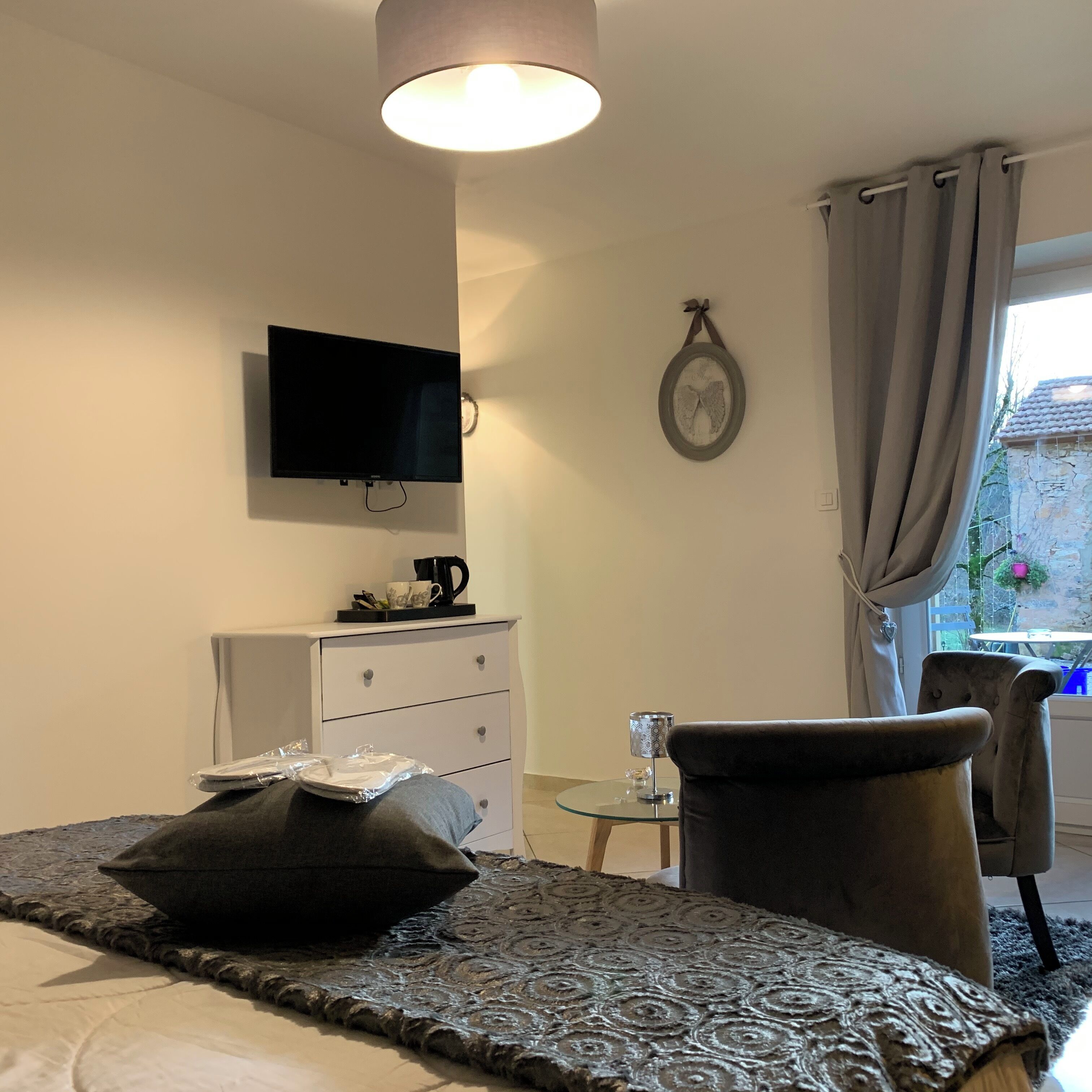 A hotel room with a bed, a TV mounted on the wall, a dresser, a small table with a lamp, and a view of the outside through a window with curtains.