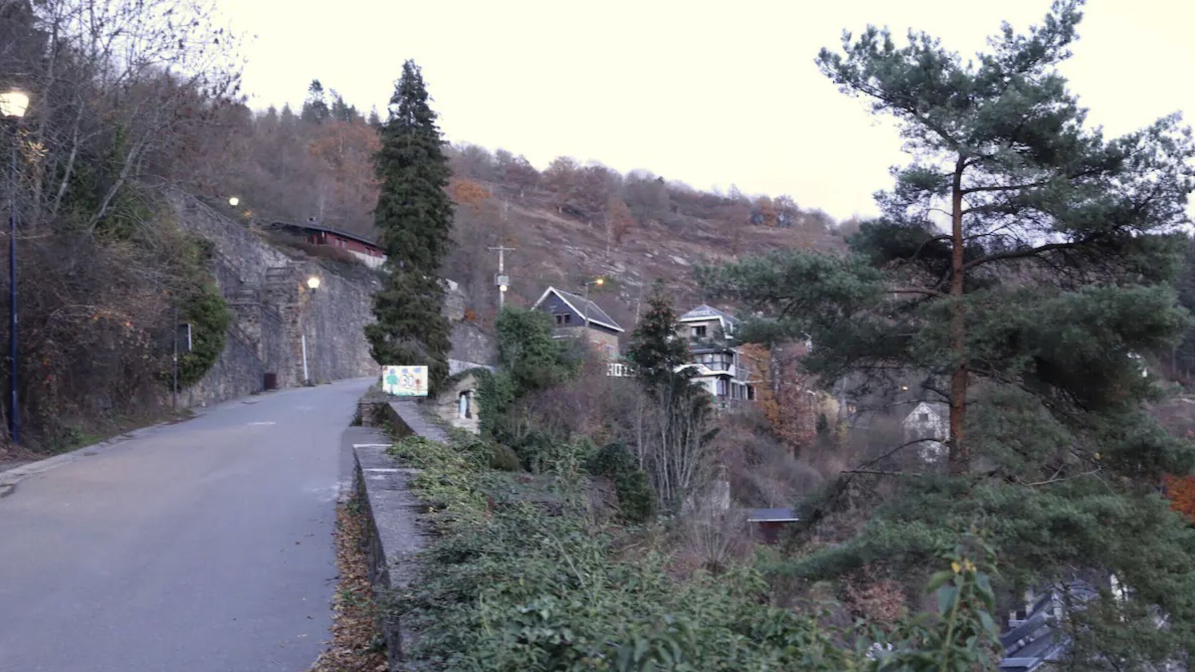 A narrow road with stone walls on one side and a forested hillside on the other.