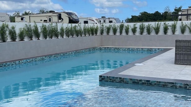Una piscina con borde de azulejos, rodeada por un muro bajo y una fila de plantas.