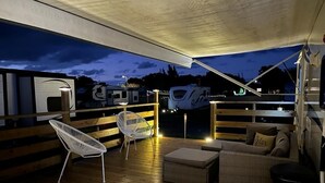 Una terraza techada al aire libre con piso de madera, una silla blanca, un sofá y una mesita.