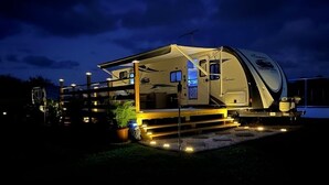 Una caravana bien iluminada con un porche cubierto y una pequeña terraza, equipada con iluminación exterior.