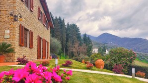 A stone building with red shutters, a well-maintained lawn, and a view of mountains in the background. - VILLA CAMELIA TUSCANY villa with pool surrounded by olive trees - wonderful view (Pescia)