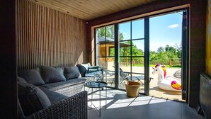 A modern living room with a sofa, a small table, and a wicker chair. There is a large glass door leading to an outdoor area with a wooden deck and a colorful unicorn decoration.