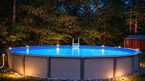 Piscina retangular acima do solo com escada, cercada por um revestimento de pedra, situada em área gramada com árvores ao fundo.