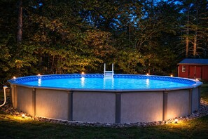 En rektangulær pool over jorden med en stige, omgivet af en stenkant, placeret i en græsplæne med træer i baggrunden.
