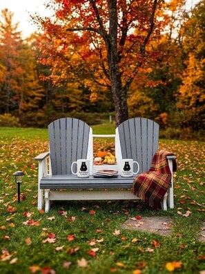 A bench with two chairs, a mug, and a blanket in a park with autumn leaves.