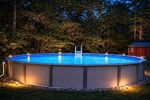 A circular above-ground pool with a ladder, surrounded by a stone border, set in a grassy area with trees in the background.