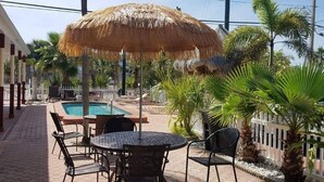 An outdoor pool area with a round table and chairs, surrounded by palm trees and a thatched umbrella.