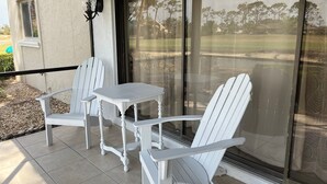 A patio with a white table and two white chairs, a black lantern, and a view of a golf course through a glass door.