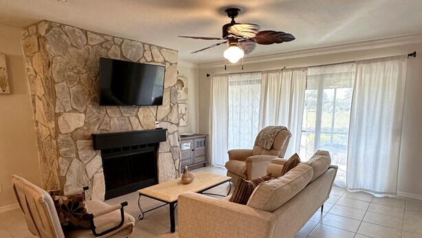 A living room with a stone fireplace, a flat-screen TV, a ceiling fan, and a large window with curtains.