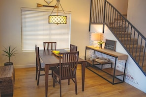 A dining area with a wooden table and chairs, a hanging light fixture, and a staircase with a metal railing.
