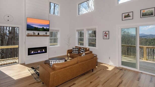 A spacious living room with a large TV, a fireplace, a brown sofa, and large windows offering views of the outdoors.