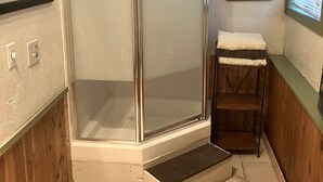 Un baño pequeño con cabina de ducha de vidrio, un banco de madera, un estante pequeño y dos cuadros en la pared.