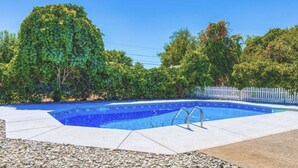 Piscina retangular com escada de metal, cercada por grade branca e vegetação exuberante.