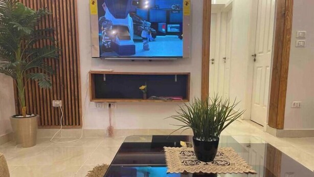 A living room with a flat-screen TV mounted on the wall, a glass coffee table, a potted plant, and a wooden floor. - 3 bedrooms Family apartment (محافظة القاهرة‬)
