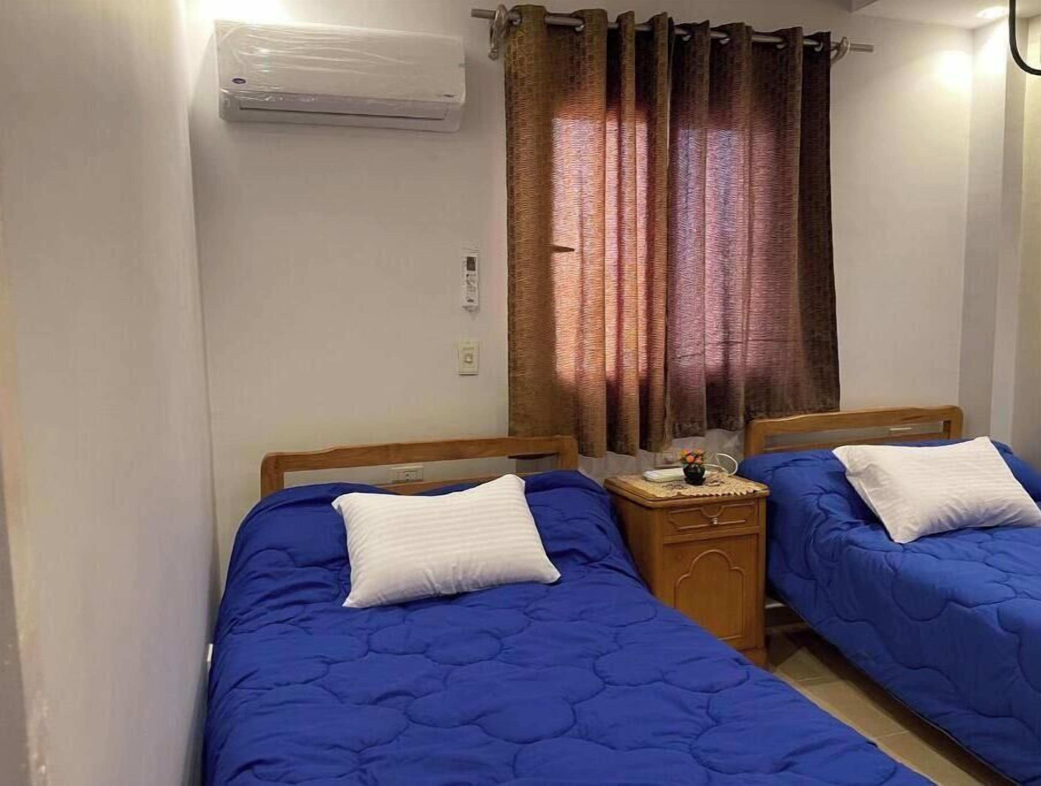 Quarto com duas camas, cada uma com colchão matelado azul e almofadas brancas. Tem uma mesa de cabeceira de madeira com uma pequena mesa e um vaso de flores. Um ar condicionado está fixado na parede acima das camas.