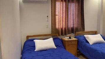 Quarto com duas camas, cada uma com colchão matelado azul e almofadas brancas. Tem uma mesa de cabeceira de madeira com uma pequena mesa e um vaso de flores. Um ar condicionado está fixado na parede acima das camas.