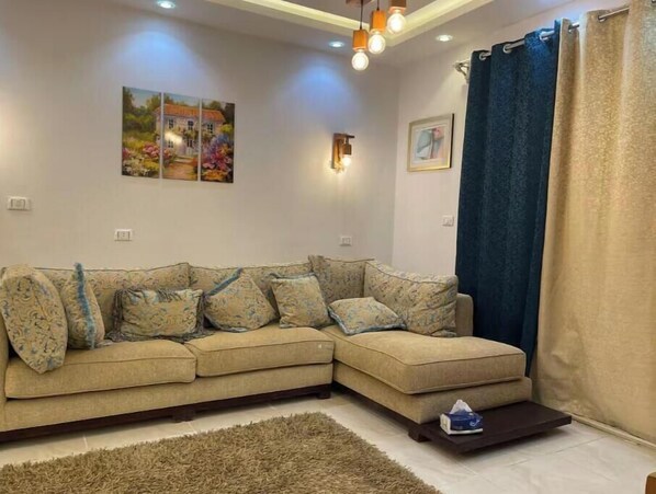 Un salon avec un canapé d’angle beige, un tapis à longues mèches et une petite table sur laquelle se trouve une boîte de mouchoirs.