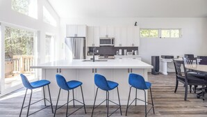 A modern kitchen with white cabinetry, a central island with bar stools, and a dining area with a table and chairs. - Luxury oasis in the woods, less than five minutes from the beach! (Wiarton)