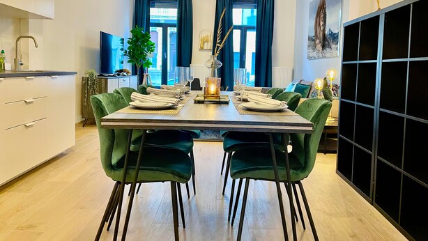 A modern dining area with a wooden table set for four, green upholstered chairs, and a view of the city through large windows. -  One Lux Stay near Fashion District (Antwerp)