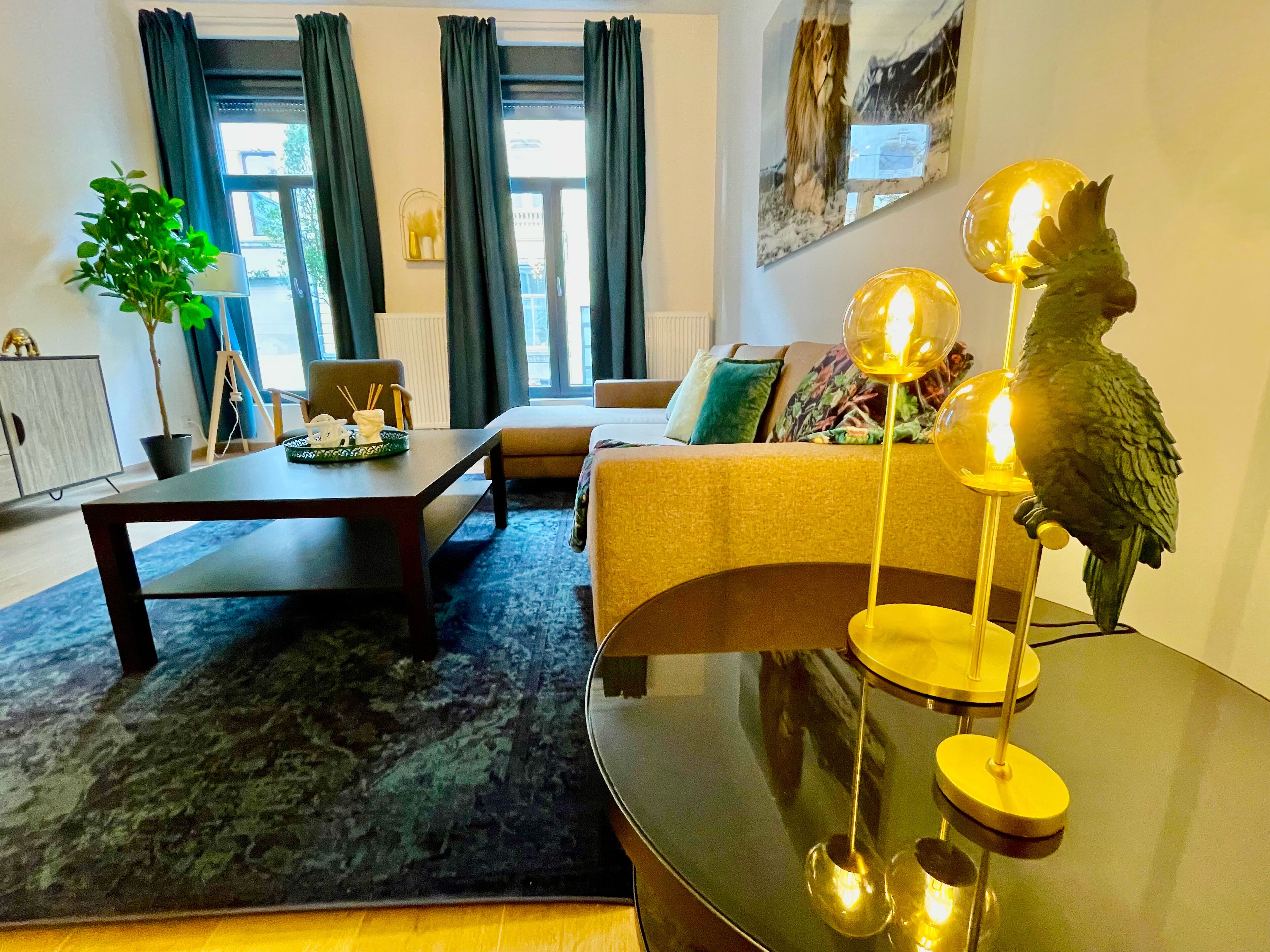 Een moderne woonkamer met een bank, een salontafel en een eettafel. Er staat een potplant en een lamp met een sculptuur van een vogel.