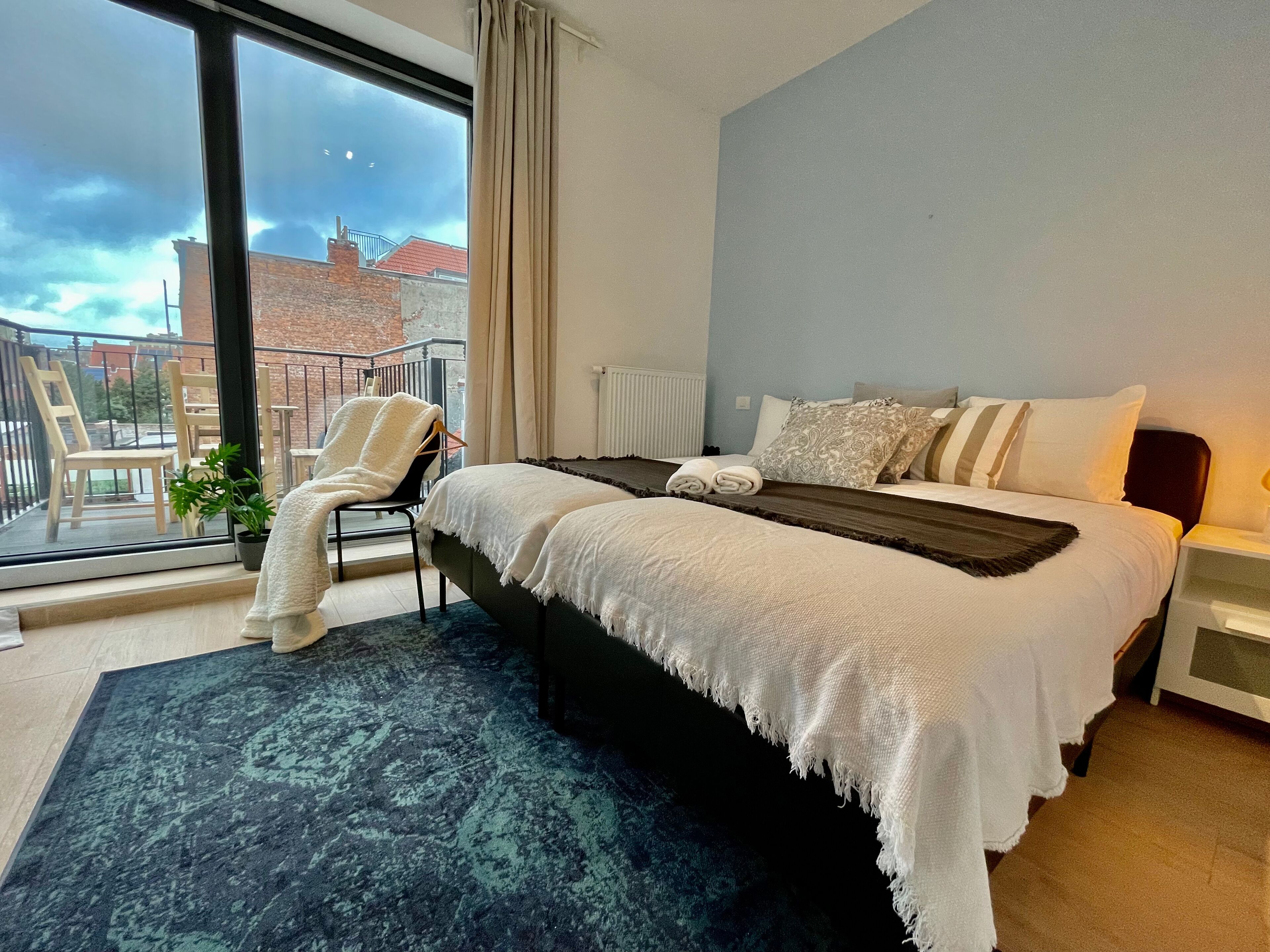 A modern bedroom with a large bed, a view of the city, and a balcony with chairs.