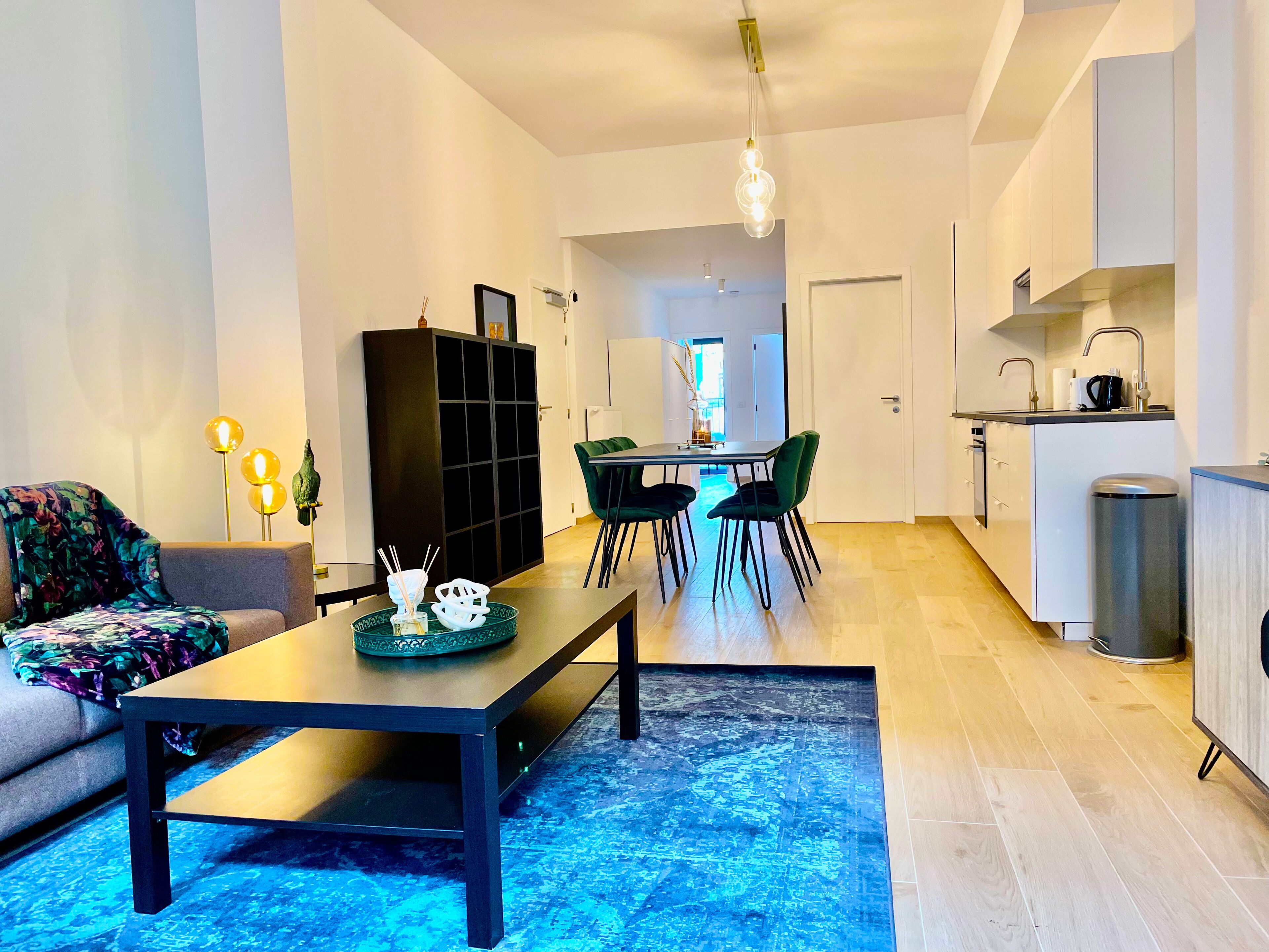 Een moderne woonkamer met een bank, een salontafel en een eethoek met stoelen en een tafel. Er is een keuken met kasten en een spoelbak.