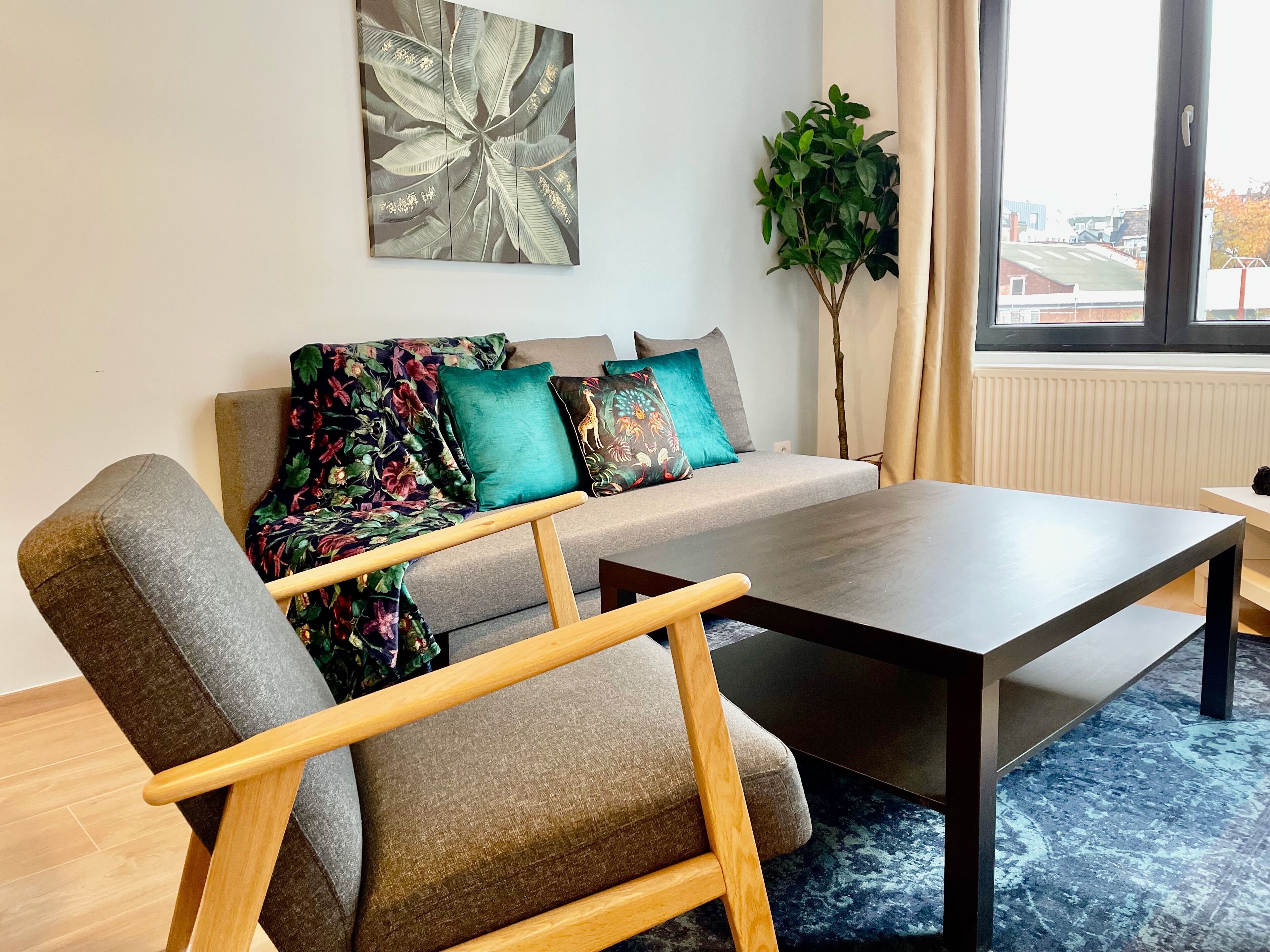 Una sala de estar moderna con un sofá gris, cojines coloridos, una mesa de centro de madera y un ventanal con vistas a edificios.