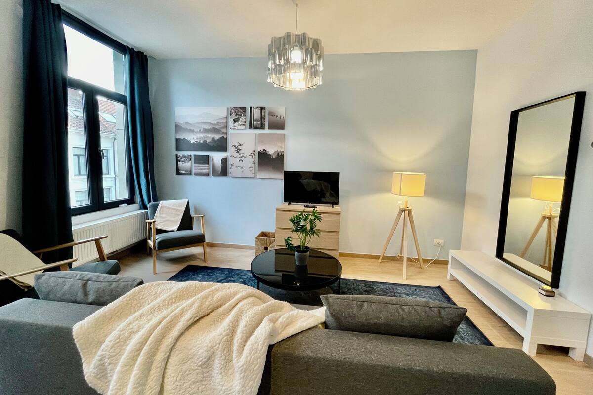Ein modernes Wohnzimmer mit einer Couch, einem Sessel und einem Couchtisch. An der Wand befindet sich ein Fernseher, eine Lampe und ein großer Spiegel.