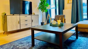 A modern living room with a flat-screen TV, a wooden cabinet, a potted plant, a table with decorative items, and a view of the outdoors through large windows. -  One Lux Stay near Fashion District (Antwerp)