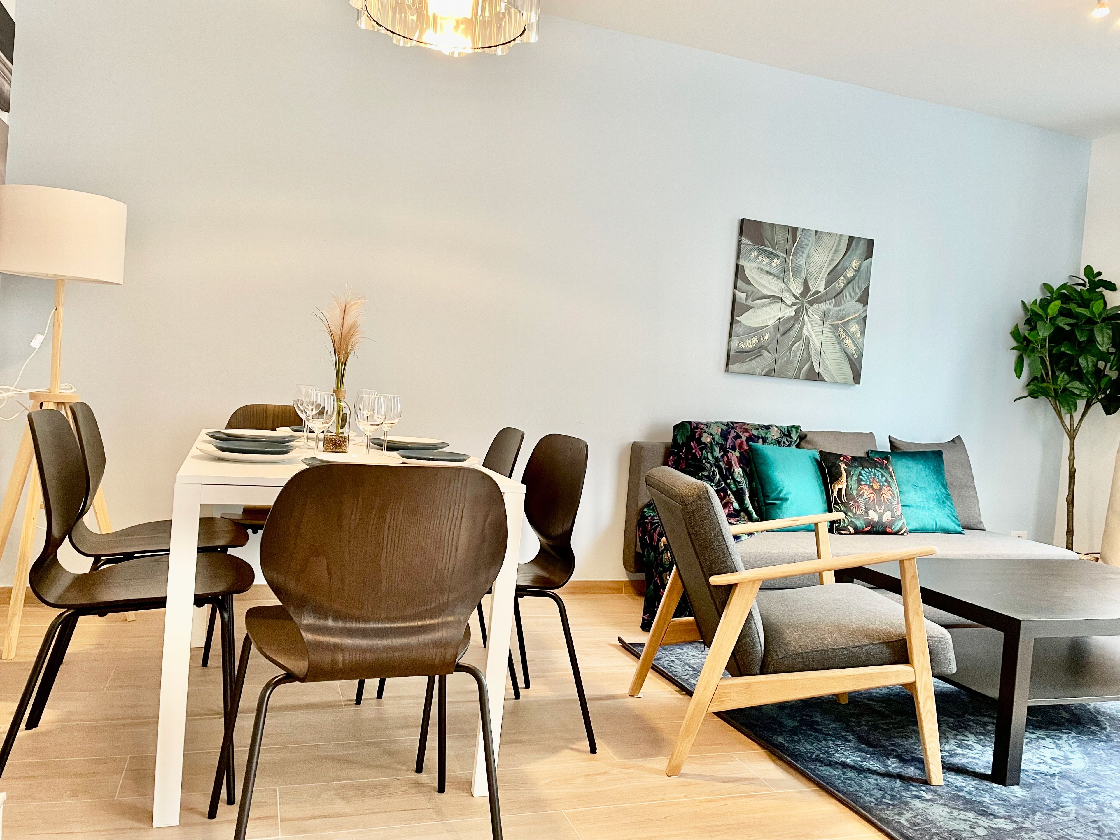A modern dining area with a white table set for four, surrounded by dark chairs. A sofa with colorful cushions is against the wall, and a large abstract painting is above it.