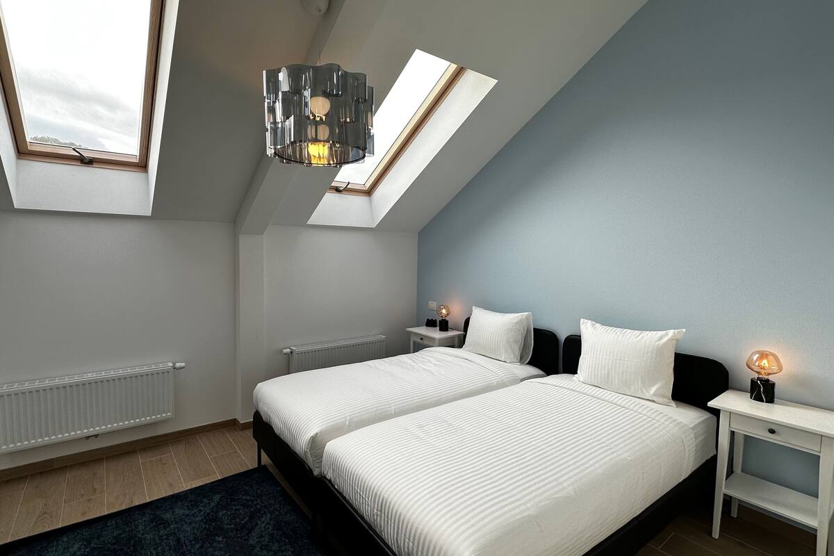 Ein Schlafzimmer mit zwei Betten, Oberlichtern, einem Heizkörper, einem Nachttisch mit Lampe und einem dunkelblauen Teppich.