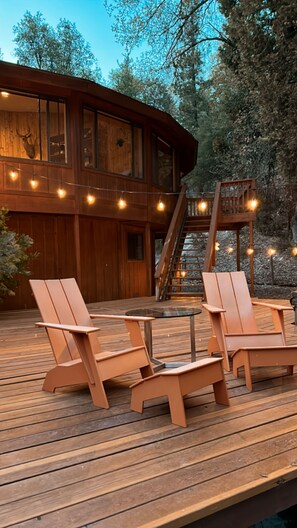 Een houten terras met ligstoelen en een glazen tafel, omringd door een houten blokhut met een trap en sfeerverlichting.