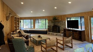 A cozy living room with a fireplace, wooden walls, and large windows. - Spacious Secluded Cabin on 9 acres Hot Tub Outdoor Dining Private Trails (Idyllwild-Pine Cove)