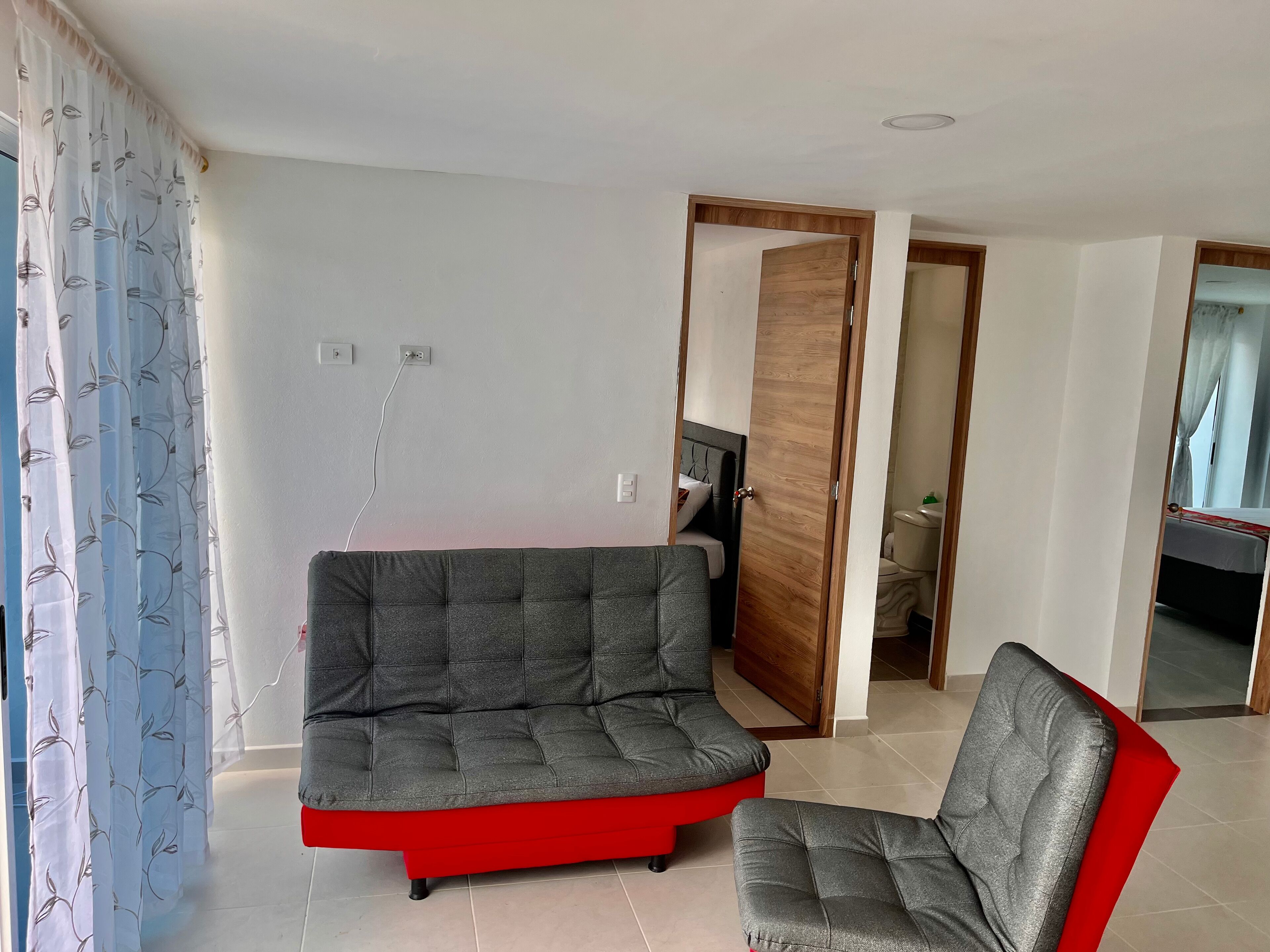 A modern living room with a grey sectional sofa and a red accent piece, a wooden door leading to a bathroom, and a tiled floor.