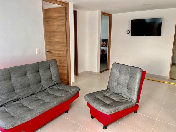 Un salon avec deux canapés gris dotés de pieds rouges, une porte en bois, un téléviseur fixé au mur et un sol carrelé.