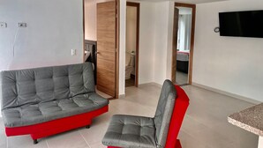 En moderne stue med en grå sofa og rød farveaccent, et fjernsyn monteret på væggen og et flisegulv.